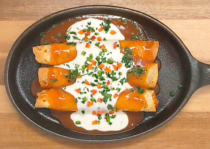 Enchiladas In A Skillet On A Wood Surface With Red Sauce, Sour Cream, Peppers And Cilantro