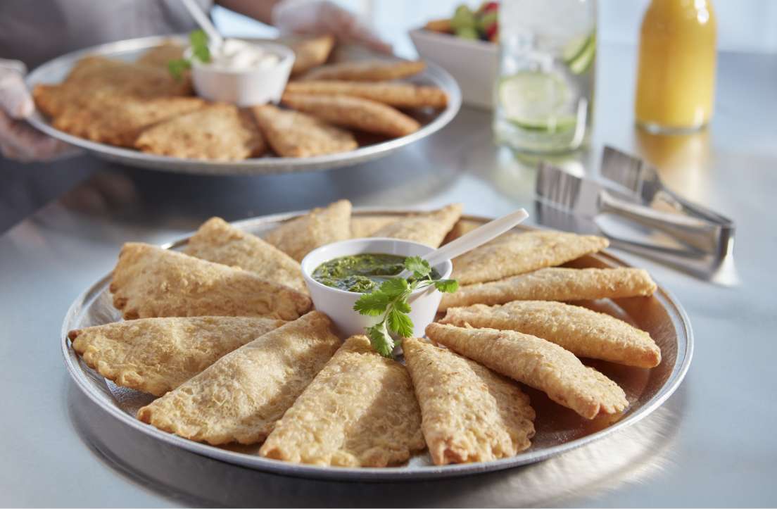 stainless steel surface with trays of empanadas with green dipping sauce in middle