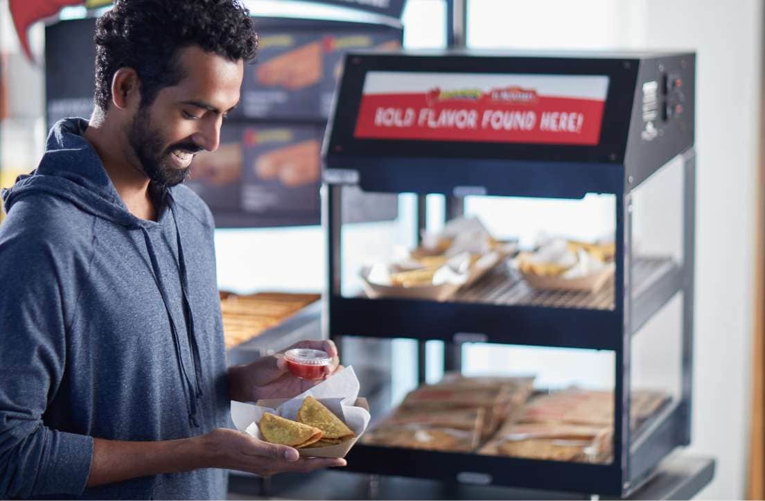 man in a blue shirt holding a paper tray of two beef mini tacos and a side of salsa in front of a convenience store hot case