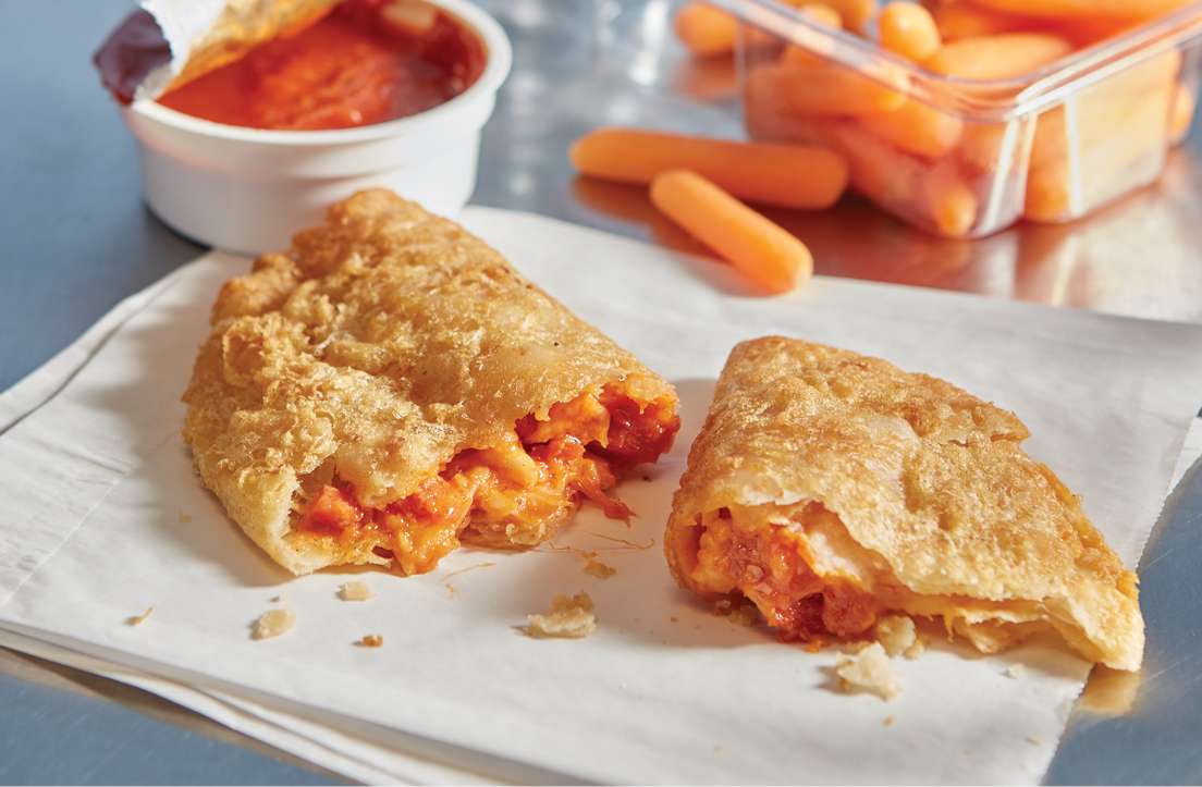 side view of one cheesy pepperoni panada broken open to show cheese and pepperoni filling on top of a white napkin with an open container of marinara sauce and container of carrots in background