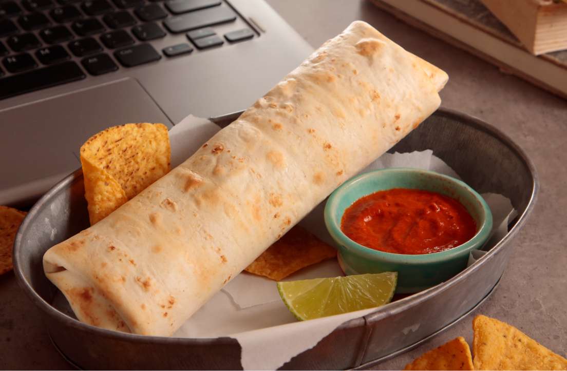 El Monterey Burrito in a tin basket lined with paper, a lime wedge, tortilla chips, and dish of salsa next to a laptop