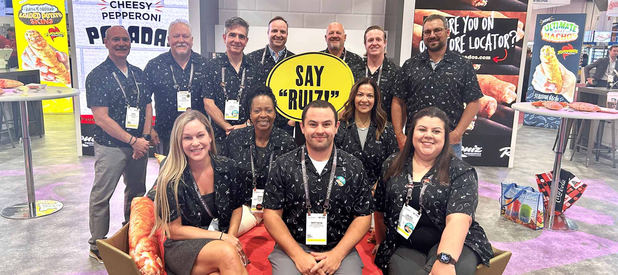 12 Ruiz Foodservice employees sitting and standing at the front of their 2024 NACS Show booth