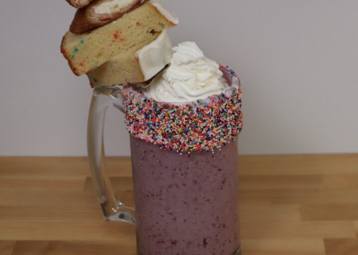 Mixed Berry Shake In A Glass Mug, Topped With Sprinkles, Whipped Creme, Fruit Loaf And Raspberry Cheescake Mini Chimi