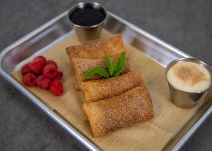 Four Raspberry Cheesecake Mini Chimis Plated On A Metal Tray With Paper, Garnished With Mint, Next To Raspberries And Two Small Dipping Containers