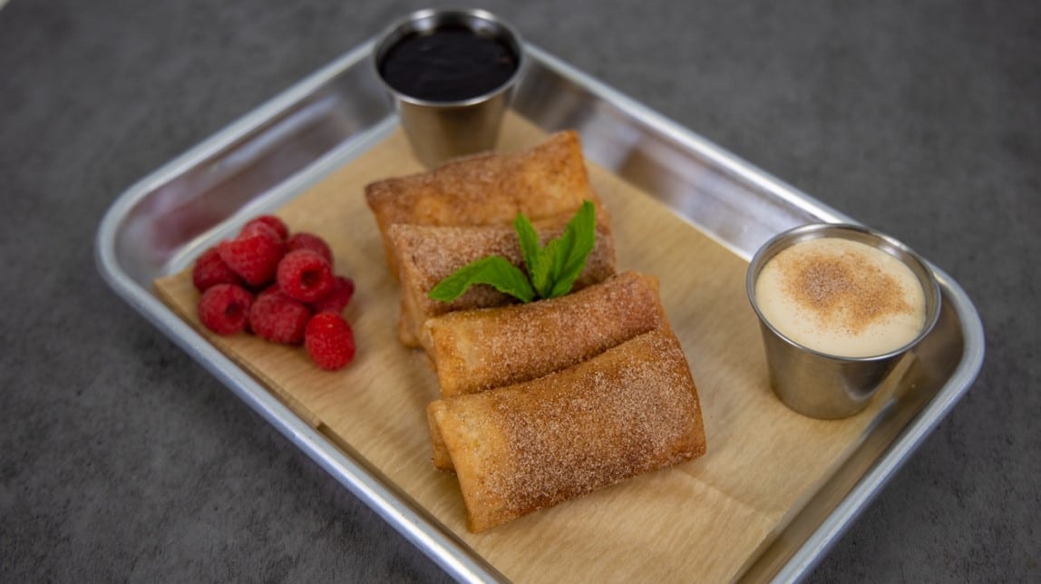 Four raspberry cheesecake mini chimis plated on a metal tray with paper, garnished with mint, next to raspberries and two small dipping containers