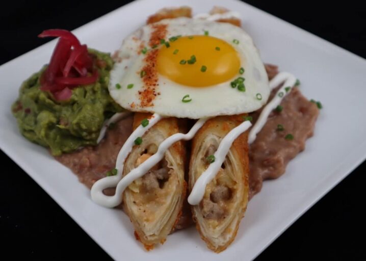 Ranchero Beef & Cheese Tornados Cut In Off Plated On Top Of Refried Beans Topped With A Fried Egg, Drizzle Of Sour Cream And Guacamole