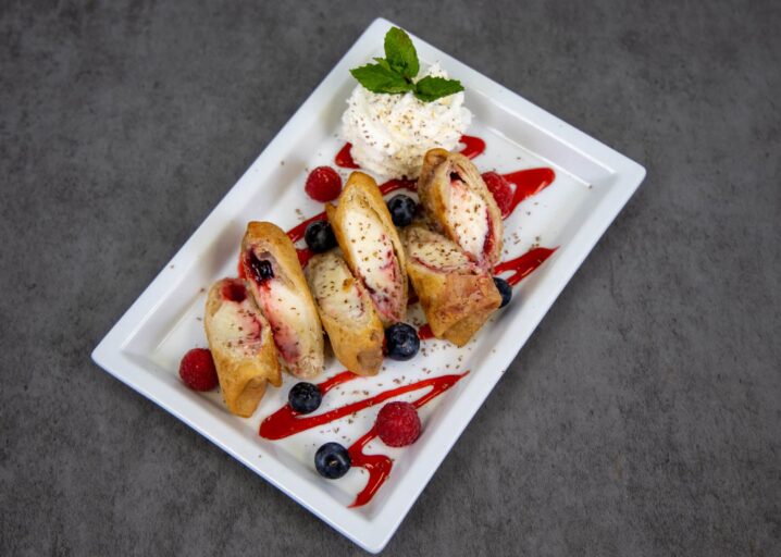 3 Sliced Raspberry Cheesecake Mini Chimis Plated And Topped With Raspberry Sauce, Raspberries, Blueberries, Whipped Cream And Garnished With Mint