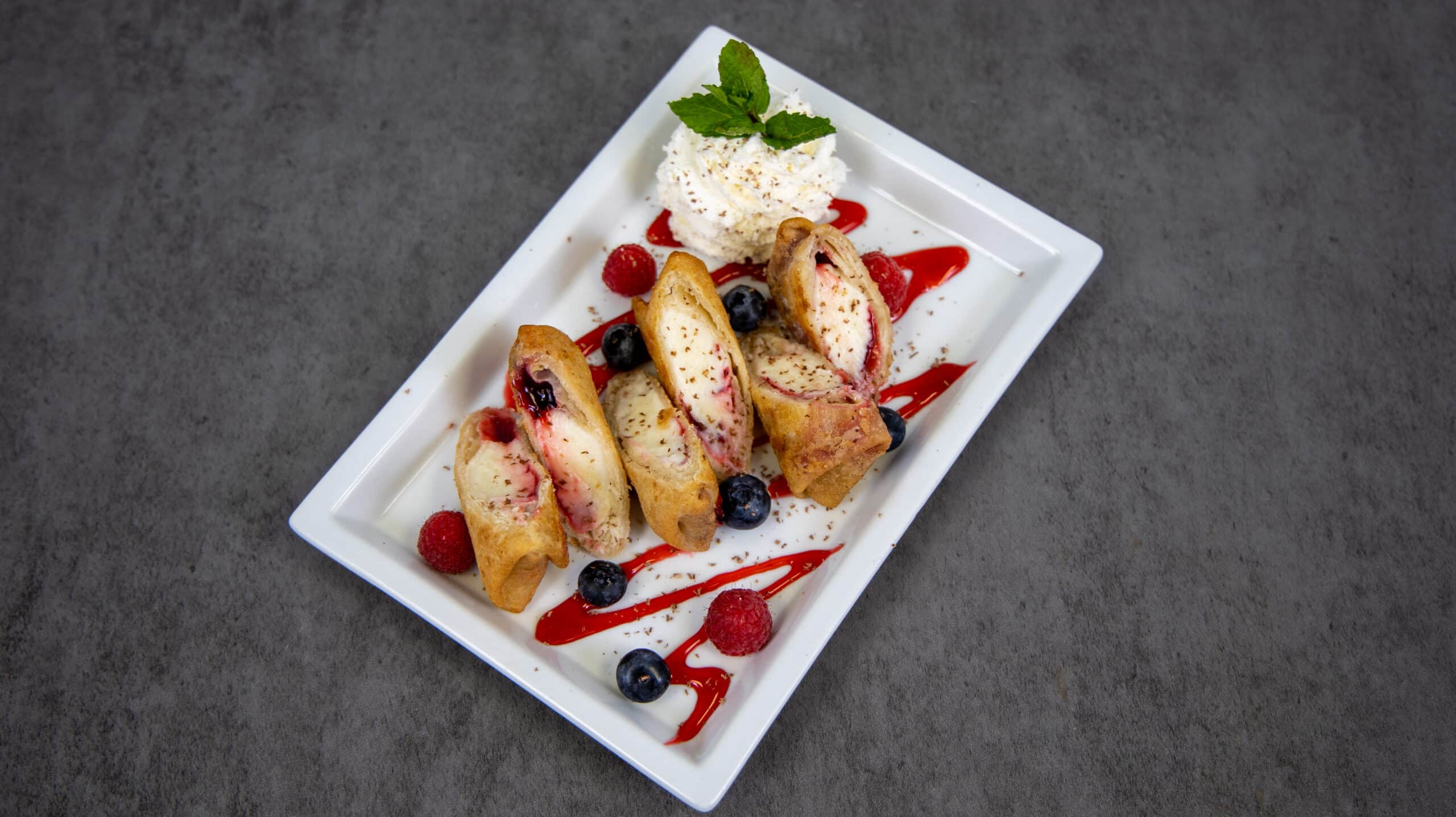 3 sliced Raspberry Cheesecake mini chimis plated and topped with raspberry sauce, raspberries, blueberries, whipped cream and garnished with mint