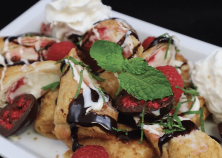 Sliced Raspberry Cheesecake Mini Chimis Plated And Topped With Chocolate Sauce, Whipped Cream, Raspberries And Mint