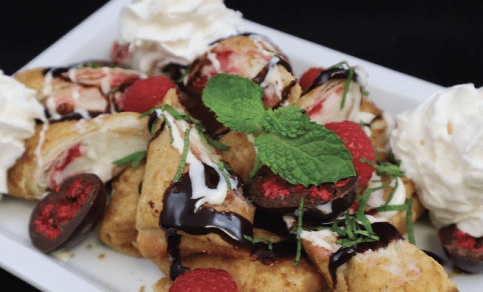 Sliced raspberry cheesecake mini chimis plated and topped with chocolate sauce, whipped cream, raspberries and mint