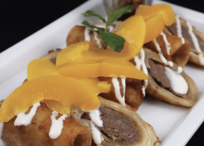 Three Sausage & French Toast Tornados Sliced And Plated, Topped With Syrup, White Chocolate Sauce And Peach Slices