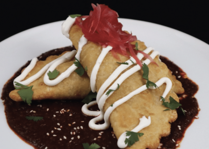 Two Bacon, Egg, Cheese & Potato Empanadas Plated On Top Of Mole Negro And Topped With Sour Cream, Cilantro And Pickled Onions