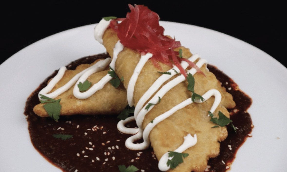 Two Bacon, Egg, Cheese & Potato Empanadas plated on top of Mole Negro and topped with sour cream, cilantro and pickled onions