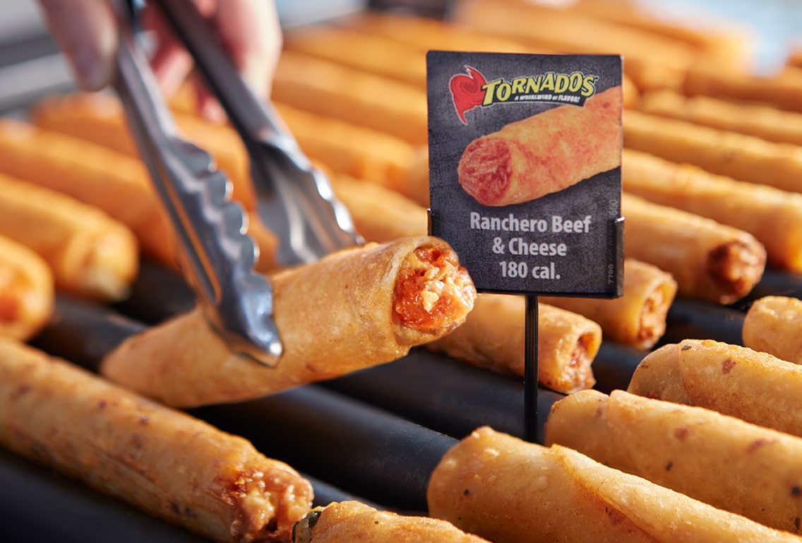 A Ranchero Beef & Cheese being grabbed from the roller grill with a pair of metal tongs