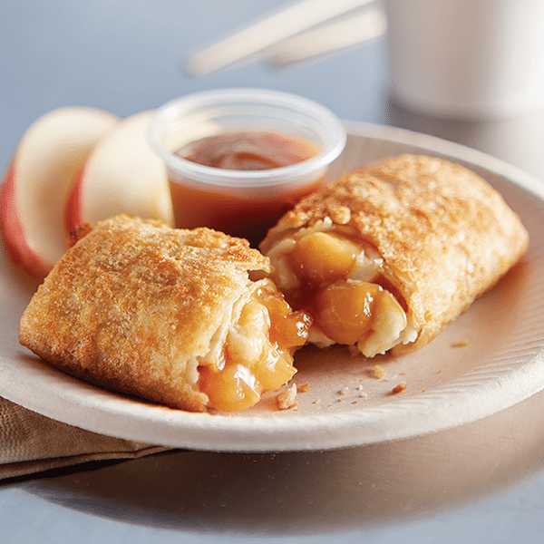 El Monterey Caramel Apple Cheesecake Mini Chimi cut open on a paper plate, showing the filliing next two a couple apple slices and dish of caramel sauce