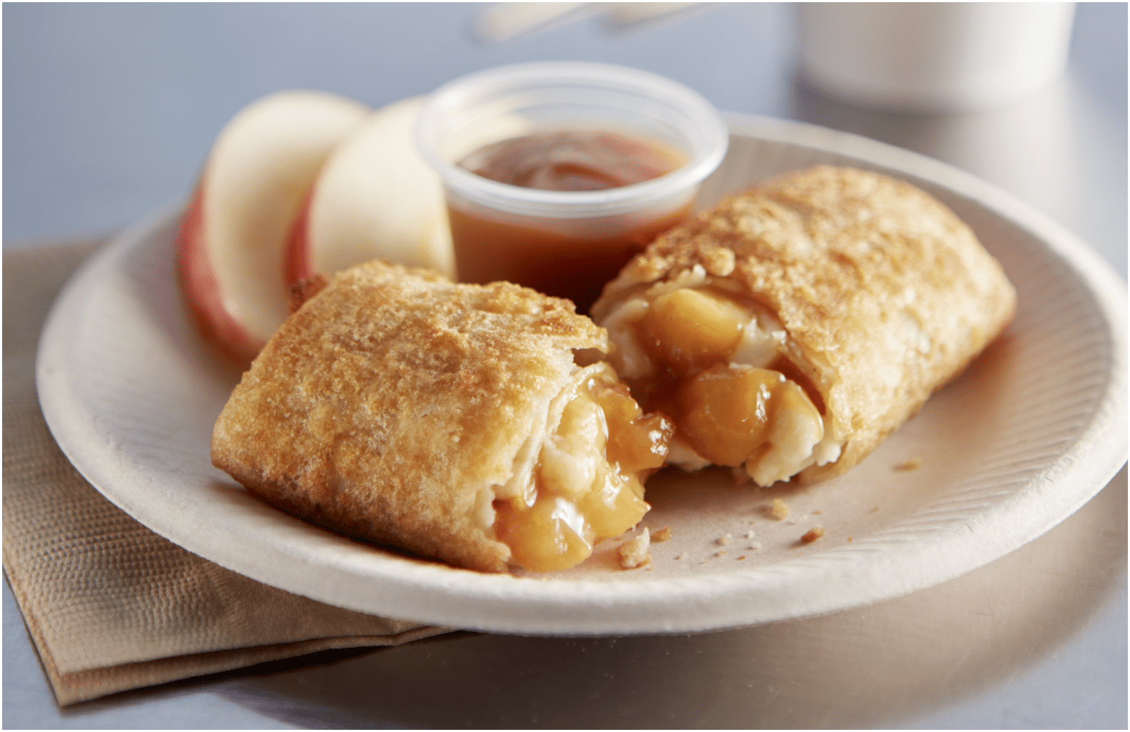 El Monterey Caramel Apple Cheesecake Mini Chimis split open showing filling on a plate with a dish of caramel and two apple slices