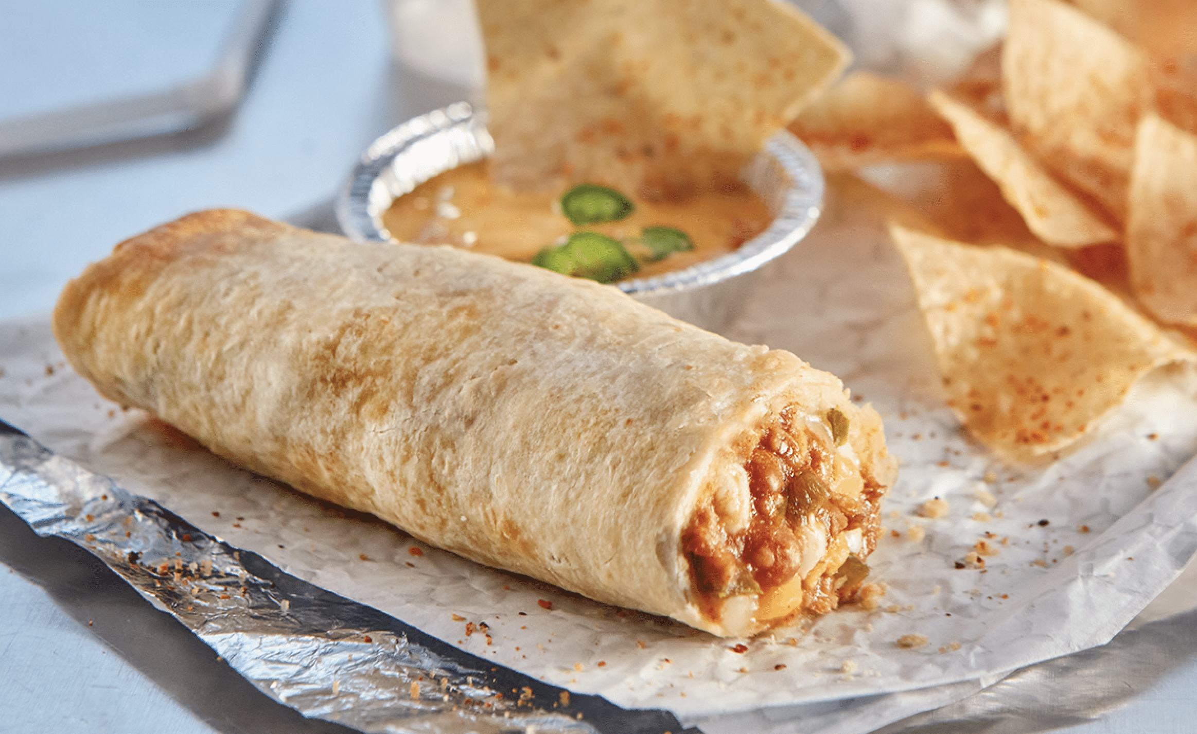 Beef, bean & cheese chimi on a foil wrap next to chips and a container of queso